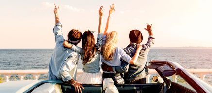 Two men and two women leaning on a car with hands raised, overlooking the ocean