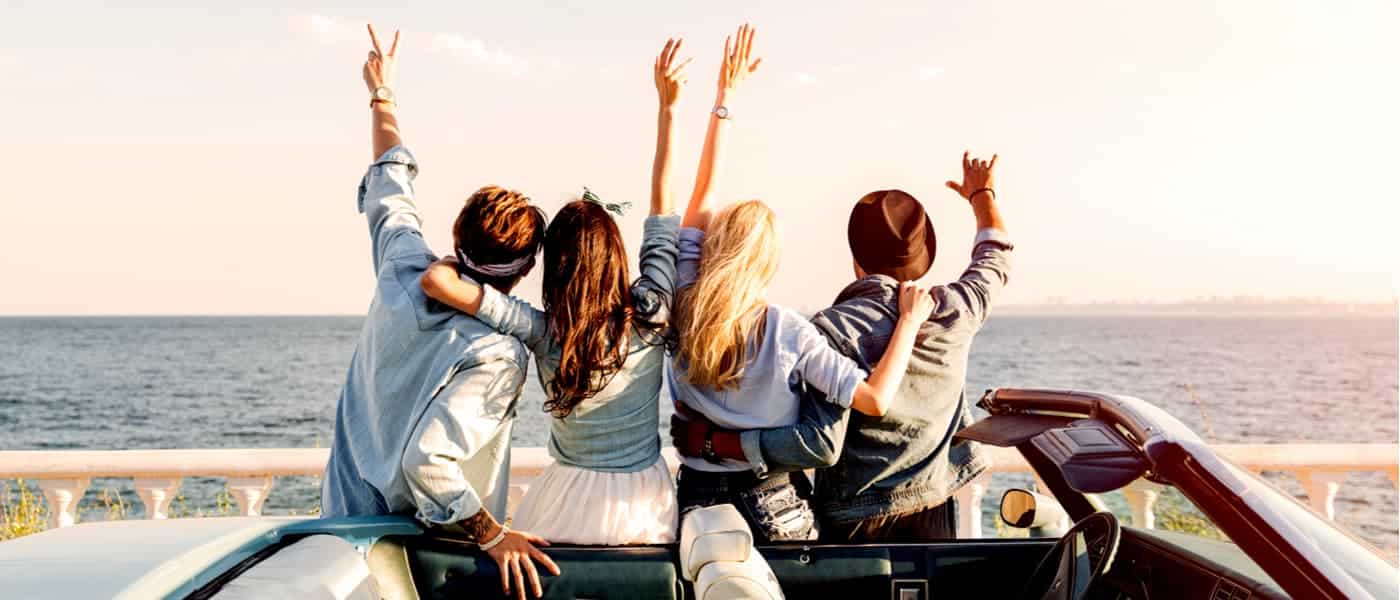 Two men and two women leaning on a car with hands raised, overlooking the ocean